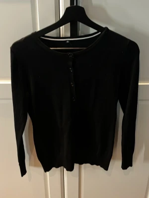 Svart kofta med knappar  - En klassisk svart kofta med rund halsringning och knäppning framtill. Långärmad och i ett mjukt, tunt material som passar perfekt till lager-på-lager-stil. Enkel och stilren design som funkar till det mesta. Aldrig använd 