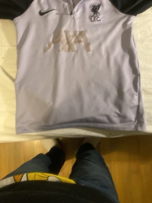 Ljuslila Liverpool träningströja Nike - Snygg ljuslila träningströja från Nike med Liverpool FC-logga och AXA sponsortryck på bröstet. Tröjan har svarta ärmar, halv dragkedja och slim fit-passform. Perfekt för träning eller att bära till vardags. Tillverkad i polyester. Köpt för 500 kr