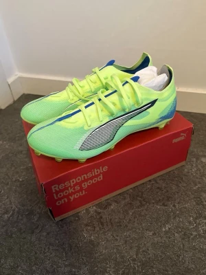 Neongröna Puma fotbollsskor - Snygga neongröna fotbollsskor från Puma med svart och silverfärgad logga på sidan. Skorna har blå detaljer och är tillverkade i lätt syntetmaterial för bästa känsla på planen. Perfekta för dig som vill sticka ut och ha bra grepp på gräs.