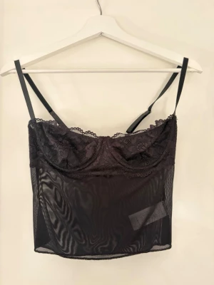 Svart spetsbody med meshdetaljer - Snygg svart body med tunna justerbara axelband, spets upptill och meshmaterial nedtill. Kuporna har bygel och spetsdetaljer, ryggen har en liten öppning och knäppning. Perfekt för dig som gillar en trendig och stilren look.