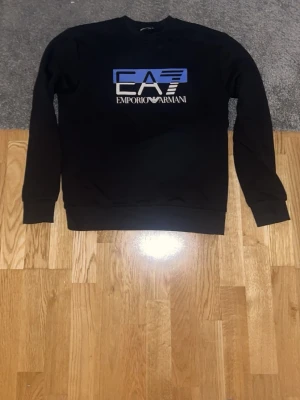 Svart sweatshirt från Emporio Armani - Svart sweatshirt från Emporio Armani EA7 med tryckt logga i blått och vitt på bröstet. Tröjan har rund halsringning, långa ärmar och ribbade muddar vid ärmslut och nederkant. Perfekt för en stilren och sportig look.