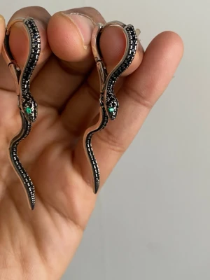 s925 sterling Silvriga ormörhängen med svarta stenar - Unika örhängen i form av slingrande ormar i silverfärg med svarta små stenar längs kroppen och gröna detaljer vid huvudet. Snygg och edgy design som verkligen sticker ut. Perfekta för dig som gillar statement-smycken och vill ha något annorlunda. material: s925 sterling silver pläterat 