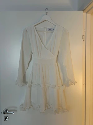 Vit omlottklänning från Dennis Maglic - Superfin vit kort omlottklänning från Dennis Maglic med långa ärmar och volangdetaljer vid ärmslut och kjolkant. Klänningen har en markerad midja och dragkedja i ryggen. Perfekt för sommarens alla tillfällen!