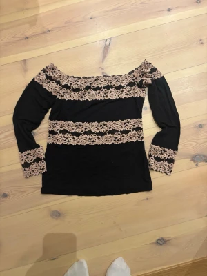 Svart offshoulder topp med spets - Snygg svart offshoulder topp med beige spetsdetaljer längs halsringning, ärmslut och över bröstet. Långärmad modell som ger en trendig och romantisk look. Perfekt att styla med jeans eller kjol. Från intimissimi 