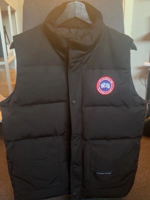 Canada Goose - Dunväst - Svart - Svart dunväst, Canada Goose med hög krage, två stora framfickor och klassisk sydd logotyp på bröstet. Västen har tryckknappar framtill och är tillverkad i slitstarkt material med dunfyllning. Använd ca 2-3 gånger och sedan bara hängt i garderoben. Den är i nyskick och har inga tecken på användning.