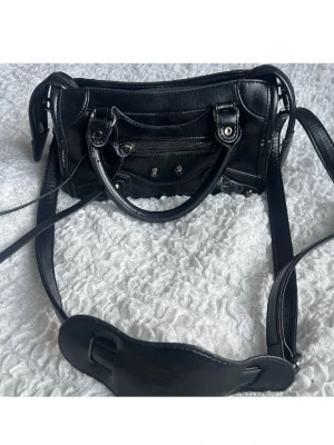 Svart  handväska i läder - Säljer en svart handväska i mjukt läder. Väskan har både handtag och lång axelrem, silverfärgade detaljer, dragkedja framtill och dekorativa nitar. Perfekt för dig som gillar en edgy och trendig look.