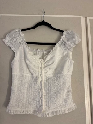 Vit topp med spets och puffärm - Superfin vit topp med spetsdetaljer, puffärmar och knappar framtill. Blusen har volangkant nertill och v-ringning. 