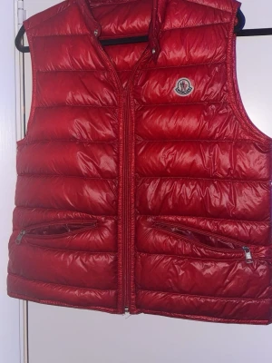Röd dunväst från Moncler - Snygg röd dunväst från Moncler med glansig finish och klassisk quiltad design. Västen har två dragkedjefickor framtill och Moncler-logga på bröstet. Perfekt för lager-på-lager och ger en cool streetstyle-vibe.