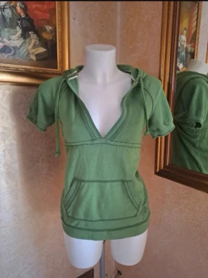 Grön hoodie med kort ärm från Abercrombie - Snygg grön hoodie från Abercrombie & Fitch med korta ärmar, djup V-ringning och huva med dragsko. Stor magficka framtill och broderad logga nere vid fållen. Tillverkad i mjuk bomull, perfekt för en avslappnad stil. 