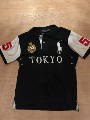 Ralph Lauren tokyo - Svart t-shirt från Polo Ralph Lauren med vita ärmar och stora röda siffror 5 på ärmar och rygg. Framsidan har broderad logga, Tokyo-tryck och emblem. Klassisk pikékrage och knappslå framtill.