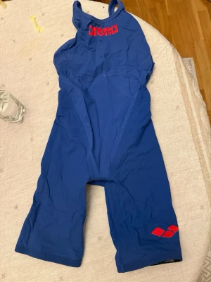 Blå tävlingsbaddräkt från Arena - Säljer en blå tävlingsbaddräkt från Arena med röd logga framtill och röda detaljer. Dräkten har långa ben och breda axelband för extra stöd. Insidan har ett svartvitt geometriskt mönster och röda korsade band. Perfekt för simträning eller tävling.