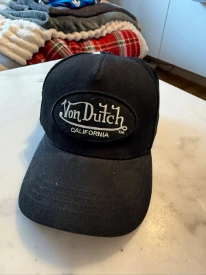 Svart Von Dutch truckerkeps - Säljer en svart truckerkeps från Von Dutch med klassisk broderad logga framtill och texten 'California'. Kepsen har justerbar rem bak och meshpaneler för extra ventilation. Perfekt för dig som gillar streetwear och vill ha en ikonisk accessoar.