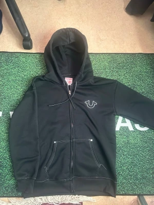 Svart hoodie från True Religion - Säljer en svart hoodie från True Religion med dragkedja och vit logga broderad på bröstet. Hoodien har vita kontrastsömmar, två fickor fram och justerbar huva med snören. Perfekt för en chill och stilren look.