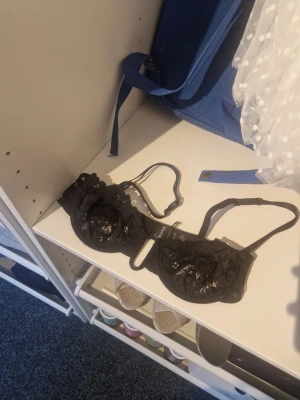 Svart spets-bh med paljettdetaljer - Snygg svart bh med genomskinlig spets och glittriga paljettdetaljer. Bh:n har justerbara axelband och dekorativa kanter. Perfekt för dig som gillar en elegant och trendig look. Aldrig använd