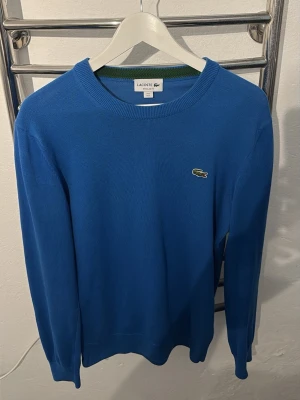 Blå stickad tröja från Lacoste - Snygg blå stickad tröja från Lacoste med klassisk rund halsringning och den ikoniska krokodilloggan broderad på bröstet. Tröjan har långa ärmar och ribbade muddar vid ärmslut och nederkant. Perfekt för dig som gillar stilren och enkel design.