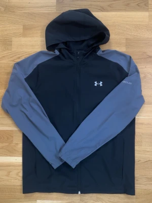 Under Armour svart och grå vindjacka - Snygg vindjacka från Under Armour i svart med grå ärmar och huva. Jackan har dragkedja framtill, elastiska muddar och logga på bröstet. Perfekt för dig som gillar sportig stil och vill ha något lätt och smidigt med huva.