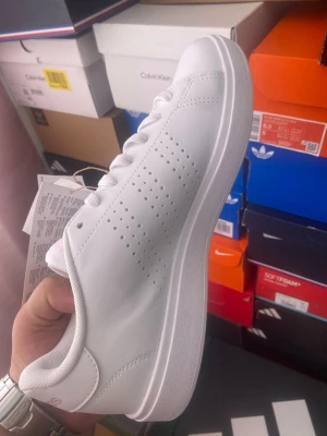 Vita Adidas sneakers i läder - Fräscha helvita sneakers från Adidas med klassisk låg profil. Skorna har perforerade ränder på sidorna istället för de vanliga tre ränderna och Adidas-logga baktill. Tillverkade i slätt läder med vit sula och vita skosnören. Perfekta för en clean och stilren look.