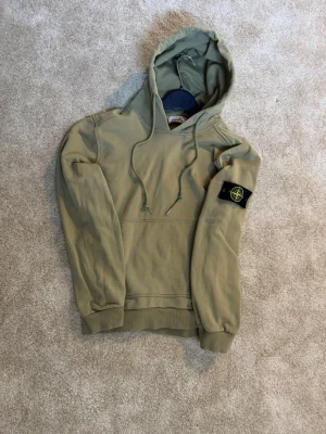 Mörkgrön hoodie från Stone Island - Snygg beige hoodie från Stone Island med klassisk logotyp-patch på vänster ärm. Hoodien har huva med dragsko, magficka och ribbade muddar. Sitter lagom tajt och inte för pösig för skön komfort och avslappnad stil.