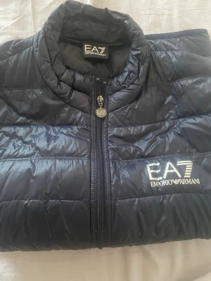 Svart dunväst från EA7 Emporio Armani - Snygg svart dunväst från EA7 Emporio Armani med quiltad design och dragkedja framtill. Västen har hög krage och tydlig logga på bröstet. Perfekt för lager-på-lager och ger en sportig look. Materialet är glansigt och lätt, vilket gör den smidig att bära.