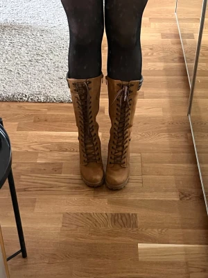 Bruna snörade höga läderstövlar - Snygga bruna stövlar i läder med högt skaft och grov klack. Stövlarna har snörning hela vägen upp och en cool vintagekänsla. Perfekta till både kjol och jeans för en trendig look. De finns lite sprickor i lädret precis ovanför klacken 