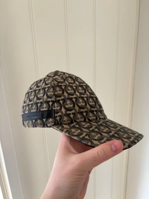 Beige keps från Salvatore Ferragamo - Snygg beige keps från Salvatore Ferragamo med svart mönster över hela kepsen och en svart läderdetalj med logga på sidan. Justerbar rem baktill. Perfekt accessoar för en trendig look.