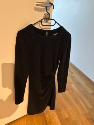 Svart glittrig kort klänning XS - Snygg svart glittrig kort klänning från Cubus i storlek XS. Klänningen har lång ärm, rund halsringning och en snygg drapering i midjan. Perfekt för dig som vill sticka ut med en elegant look.