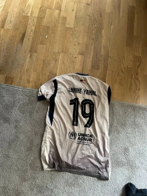 Barcelona Lamine Yamal matchtröja - Säljer en beige FC Barcelona matchtröja med Lamine Yamal och nummer 19 på ryggen. Tröjan har svarta detaljer plural, UNHCR och Spotify tantryck samt klubbmärke på bröstet. Tillverkad i lätt och vent forced material, perfekt för fotbollsträning.