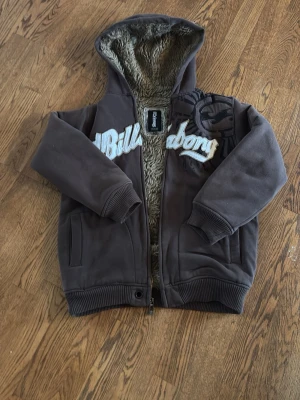 Brun sophoodie från Billabong - Mysig brun hoodie från Billabong med fluffigt foder i hela insidan och huva. Stor logga broderad över bröstet och ribbade muddar vid ärmar och nederkant. Två fickor framtill och dragkedja. Perfekt för kalla dagar och riktigt skön att ha på sig. Är dock för liten för mig