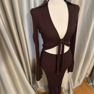 Gina Brun ribbad långärmad klänning med cut out - Trendig brun långärmad klänning med ribbad struktur och knyt framtill. Djup v-ringning och snygg cut-out vid midjan ger en modern look. Klänningen har en kroppsnära passform.