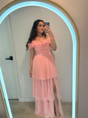 Rosa balklänning  - Romantisk ljusrosa långklänning med flera lager tyll, offshoulder-modell och stora volangdetaljer vid bysten. Klänningen har slits på ena sidan och markerad midja för en feminin siluett. Perfekt för bal eller fest. Hade denna underbart fina på min bal, och fick tusentals komplimanger!! Verkligen sjukt vacker klänning, men kommer inte ha någon nytta av den mer. Köpte denna för 2000 kr, och haft den endast en gång.