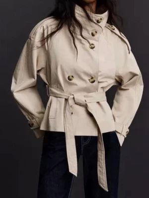 Beige kort trenchcoat med bälte - Stilren beige trenchcoat med dubbelknäppning, höga axlar och knytskärp i midjan. Jackan har bred krage, dekorativa axelklaffar och stora knappar. Perfekt för dig som vill ha en trendig och klassisk look. Den är helt ny med prislapp så därav aldrig använd. 