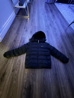 Svart pufferjacka från GANT - Snygg svart pufferjacka från GANT med huva och dragkedja framtill. Jackan har en quiltad design och två stora fickor på sidorna. Perfekt för kalla dagar när du vill hålla dig varm och stilren.