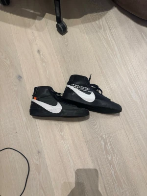 Off-White x Nike Blazer Mid svart/vit - Säljer ett par Off-White x Nike Blazer Mid sneakers i svart med vit swoosh och textdetaljer på sidan. Skorna har högt skaft, svarta skosnören och en liten orange detalj vid hälen. De är använda ganska lite, men har stått i garderoben så är ganska smutsiga. Hör av dig vid flera frågor! Priset är alltid diskuterbart.