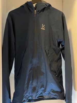 Mörkblå vindjacka från Haglöfs - Snygg mörkblå vindjacka från Haglöfs med huva och hel dragkedja framtill. Jackan har två sidofickor med dragkedja och diskret logga på bröstet. Tillverkad i lätt och slitstarkt syntetmaterial, perfekt för blåsiga dagar. 