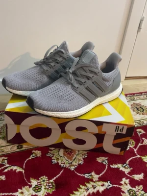 Grå Adidas Ultra Boost sneakers - Säljer ett par grå Adidas Ultra Boost sneakers med stickad ovandel och klassiska tre ränder på sidan. Skorna har vit Boost-sula för extra komfort och svart yttersula. Perfekta för dig som vill ha både stil och funktion i vardagen.