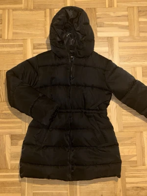 Svart pufferjacka med huva - Säljer en svart pufferjacka med huva och dragked feed. Jackan har lång ärm och en något längre passform och enkel att styla till olika outfits.