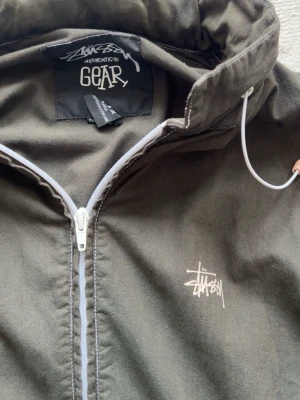Mörkgrön tunnjacka  från Stüssy - Säljer en svart tunnjacka från Stüssy med vit logotyp på bröstet och dragkedja framtill. Jackan har två fickor med dragkedja och justerbar huva med snören. Materialet är lätt och passar perfekt för våren även bra till sommar kvällar. Den är köpt i Köpenhamn och säljs för den tyvärr är lite för stor. För mer bilder/frågor så skicka dm. 