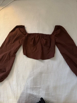 Brun croppad blus med puffärm - Säljer en brun croppad blus med bred urringning och långa puffärmar. Toppen har resår vid axlar och ärmslut för en snygg offshoulder-look. Perfekt att styla med höga jeans eller kjol för en trendig vibe.