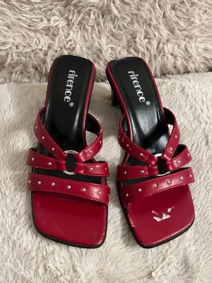 Röda kitten heels med nitar från Firence - Snygga röda kitten heels från Firence med två remmar dekorerade med små silverfärgade nitar och en rund metallring framtill. Skorna har fyrkantig tå och mellanhög klack. Materialet är konstläder med svart innersula och öppen häl.