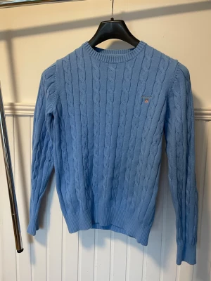 Gant Ljusblå kabelstickad tröja  - Snygg ljusblå kabelstickad tröja från Gant med rund halsringning och diskret logga på bröstet. Tröjan har långa ärmar och ribbade muddar vid ärmslut och nederkant. Perfekt för en stilren och klassisk look. Pris kan diskuteras vid snabb affär.
