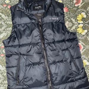 Svart dunväst från Everest - Svart dunväst från Everest med quiltad design och dragkedja framtill. Västen har två fickor med dragkedja och diskret logga på bröstet. Perfekt att slänga över en hoodie eller stickad tröja för extra värme. Snygg och enkel stil som funkar året runt.