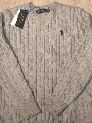 Ralph Lauren tröja  - Kabelstickad ljusgrå tröja från Polo Ralph Lauren med rund halsringning och den klassiska broderade loggan på bröstet. Tröjan har långa ärmar och ribbade muddar vid ärmslut och nederkant. Perfekt för en stilren och avslappnad look.