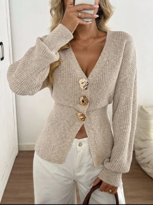 Beige ribbstickad kofta med stora knappar - Trendig beige ribbstickad kofta med djup V-ringning och tre stora, marmorerade knappar framtill. Koftan har lång ärm och figurnära passform som framhäver midjan. Perfekt att styla med höga byxor för en snygg look. Säg till om ni vill ha fler bilder💕