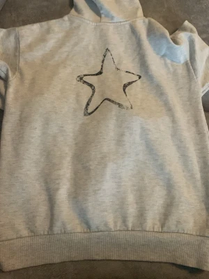 Grå hoodie med tryck 'AMOUR' - Säljer en ljusgr covering hoodie med svart text 'AMOUR' på framsidan och en stor stjärna på ryggen. Hoodien har huva och ribbade muddar vid ärmslut och nederkant. Perfekt för dig som gillar enkel men snygg streetstil.