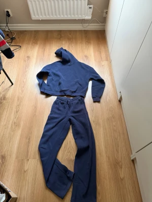 Mörkblå hoodie och byxor  - En enkel och stilren mörkblå hoodie med huva och stor magficka framtill. Hoodien har långa ärmar och ribbade muddar vid ärmslut och nederkant. Hoodie är i storlek XS och byxor XXS