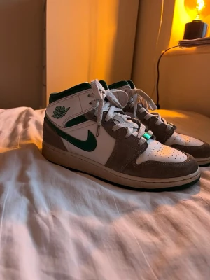 Nike Air Jordan 1 Mid vit/grön - Säljer ett par Nike Air Jordan 1 Mid sneakers i vitt och grönt med grå ”mockadetaljer”. Klassiska Jordans med gröna konturer vid snöre. Skorna har vita skosnören.