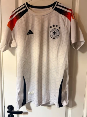 Tysklands matchtröja Adidas herr - Vit Tyskland fotbollströja från Adidas med svarta detaljer, fyra stjärnor ovanför klubbmärket och diskret mönster i tyget. Svarta, röda och orange ränder på axlarna. Perfekt för dig som älskar fotboll och vill stötta landslaget.