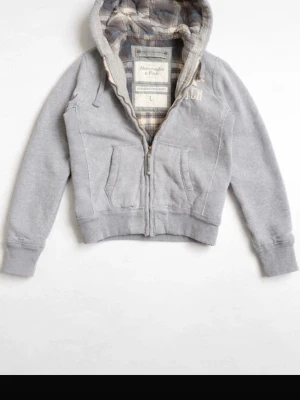 Grå hoodie från Abercrombie & Fitch - Snygg grå hoodie från Abercrombie & Fitch med dragkedja och två fickor framtill. Insidan av huvan är fodrad med rutigt tyg i grått, vitt och blått. Perfekt för en avslappnad och trendig stil. Klassisk logga broderad på bröstet.