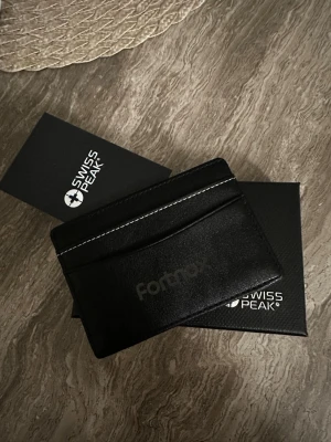 Svart korthållare från Swiss Peak - Stilren svart korthållare i läder från Swiss Peak med diskret logga och vita sömmar. Plats för flera kort på båda sidor och minimalistisk design som passar perfekt i fickan. Perfekt för dig som vill ha koll på korten utan att bära en stor plånbok. Den är stilig och fräsch. Skriv över det minsta lilla!