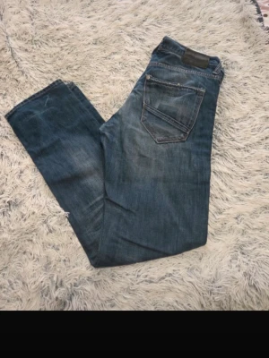 Blend jeans - Säljer ett par blå jeans från blend med raka ben och klassisk femficksmodell. Jeansen har slitningar och en liten röd detalj vid fickan fram. Tillverkade i denim med snygg tvättad look och knappgylf. Mått står i bilderna